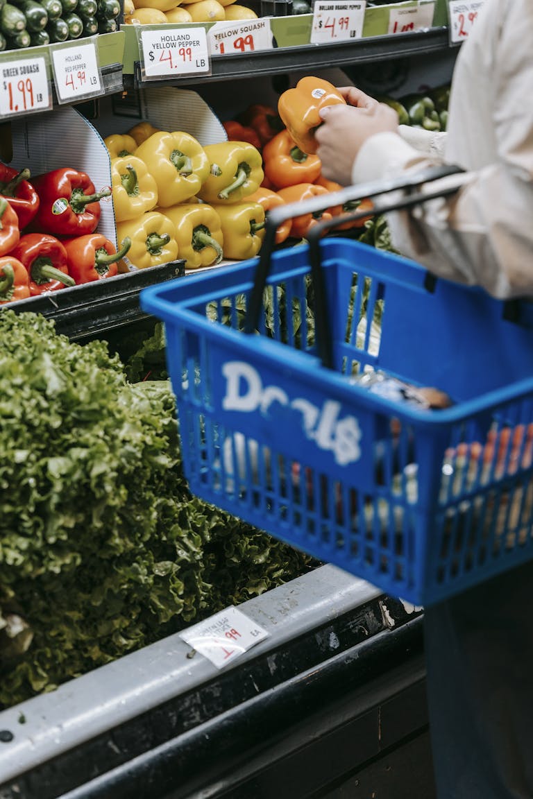 Side view of crop faceless buyer in casual clothes carrying shopping basket while choosing assorted colorful bell peppers in supermarket
